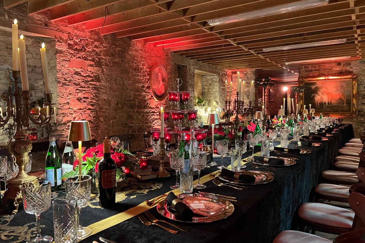 Long banquet table set for a private dinner in a candlelit stone-walled basement with red uplighting