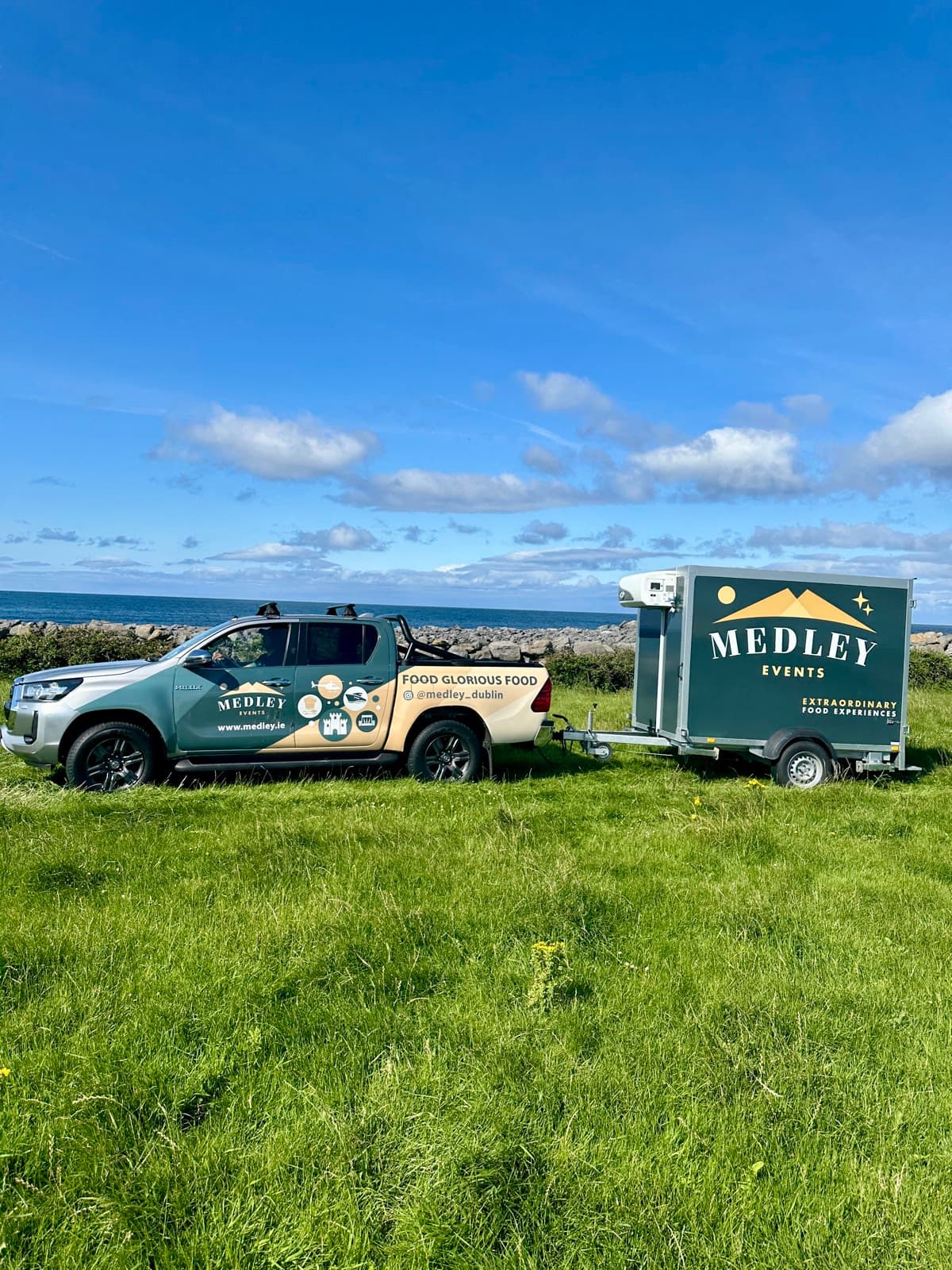 Medley's branded pickup truck and refrigerated trailer parked on a coastal grass field overlooking the sea