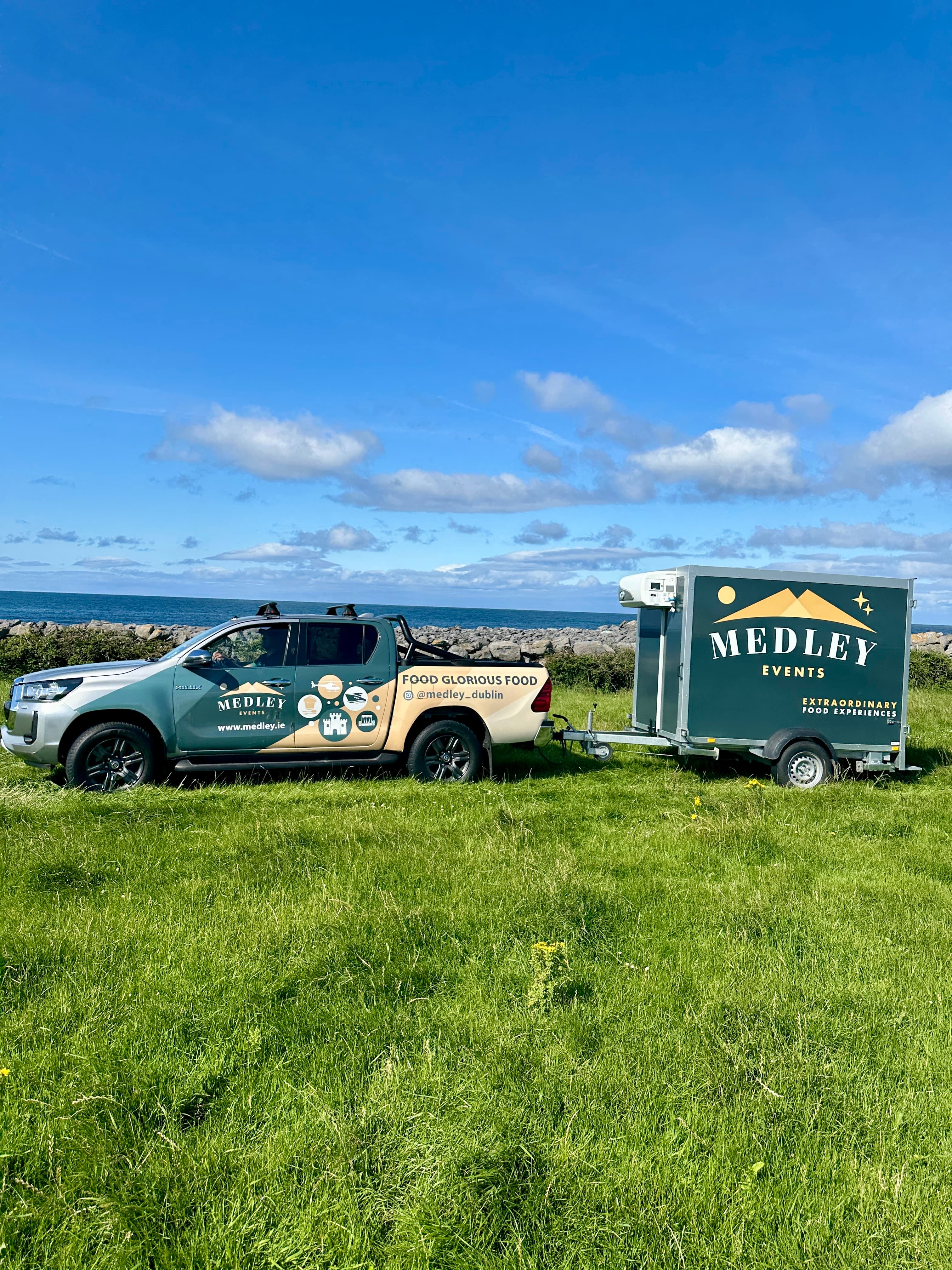 Medley's branded pickup truck and refrigerated trailer parked on a coastal grass field overlooking the sea